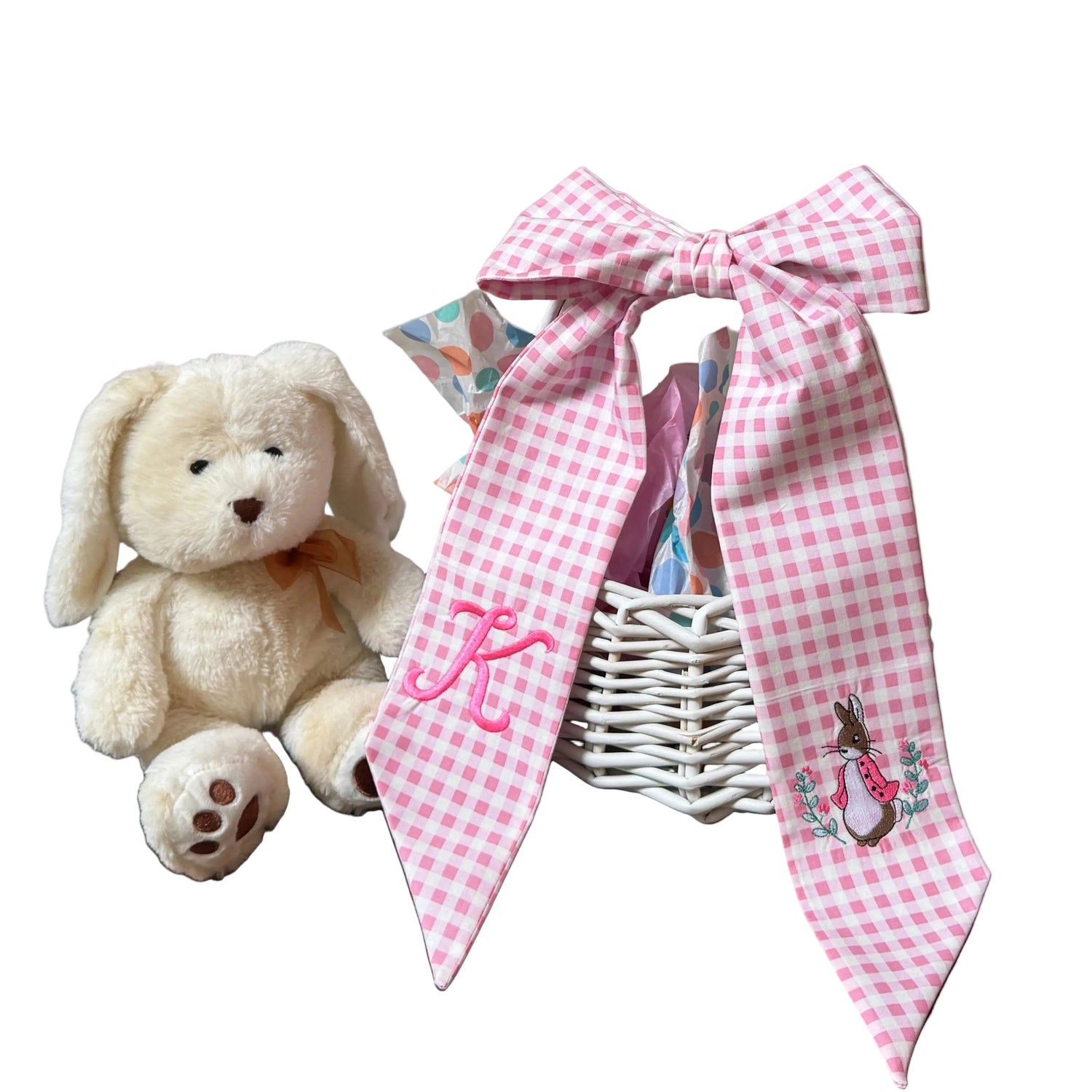 White teddy bear next to a white basket with pink gingham fabric and a rabbit design on a one sash and an initial on the other